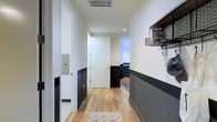 This well-appointed single-floor space makes a strong first impression with its thoughtfully designed interior. The hallway sets the tone immediately, featuring a striking two-tone wall treatment where crisp white upper walls meet a deep charcoal lower half, divided by a clean painted border. This design choice adds visual depth and a modern, polished character throughout the corridor.

Warm-toned hardwood flooring runs the length of the hallway, lending a natural warmth that contrasts beautifully with the bold wall palette. A patterned accent rug near the entry adds a welcoming touch of color and texture underfoot.

The entryway is both practical and stylish, featuring a wall-mounted wire rack shelf with hooks below — ideal for organizing everyday essentials. Recessed ceiling lighting provides even, ambient illumination throughout the corridor, while a ceiling vent points to a well-integrated HVAC system. An electrical panel is discreetly tucked behind a flush-mounted door on one side of the hallway, maintaining the clean aesthetic.

At the far end of the hallway, the space opens into a brighter area, suggesting additional rooms beyond. The overall ambiance is modern and curated, balancing industrial-inspired accents with warm, natural materials to create a cohesive and inviting environment.