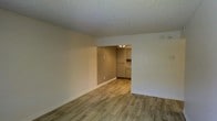 This single-floor space offers a clean, functional layout designed for comfortable everyday living. The main living area is finished with warm-toned, hardwood-style laminate flooring that runs seamlessly throughout, creating a cohesive and inviting atmosphere. Crisp white baseboards and neutral, light taupe walls provide a versatile backdrop that complements a variety of interior styles. The textured popcorn ceiling adds a classic touch overhead. An open passthrough connects the living area to the kitchen, which is outfitted with traditional raised-panel cabinetry in a warm cream finish, offering ample storage. A decorative tile backsplash adds a subtle accent to the kitchen space, and recessed lighting provides focused illumination. The open flow between the living and kitchen areas makes the space feel connected and practical. The overall ambiance is bright, neutral, and move-in ready, with a straightforward design that prioritizes functionality and ease of living.