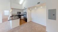 This single-floor residence is defined by its striking vaulted ceilings, which lend an airy, open feel to the main living areas. The open-concept layout seamlessly connects the living room and kitchen, where plush neutral carpeting anchors the living space and warm hardwood flooring extends through the kitchen. Ceiling fans are installed throughout, complementing the natural light that pours in through multiple windows with outdoor views of surrounding greenery.

The kitchen is well-equipped with stainless steel appliances, including a refrigerator, range, and over-the-range microwave, all set against crisp white cabinetry and dark granite-style countertops. A raised breakfast bar separates the kitchen from the living area, adding both functionality and visual interest. Track lighting and pendant fixtures provide additional illumination throughout.

The bedroom features the same soaring vaulted ceilings, plush carpeting, a ceiling fan, and multiple windows that invite ample natural light and views of the outdoors, creating a bright and comfortable retreat.

The full bathroom is finished with white cabinetry, dark granite-style countertops, a large vanity mirror with overhead lighting, and a combination soaking tub and shower. Light tile flooring and clean white walls contribute to a bright, functional space. Overall, the residence offers a cohesive, neutral palette with practical finishes throughout.