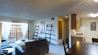 This single-floor residence offers an open-concept layout that seamlessly connects the living, dining, and kitchen areas. The living room is carpeted and features neutral walls, a textured ceiling, and a large window dressed with sheer white curtains, allowing natural light to filter into the space. Two lean-to ladder shelving units serve as functional décor accents along the main wall, complemented by framed artwork in muted tones. A gray upholstered sofa anchors the seating area, which flows naturally into the dining space. The dining area includes a wooden table and chairs, positioned near the transition to the kitchen. The kitchen is located just off the main living space and is equipped with warm wood-toned cabinetry extending to the ceiling, providing generous storage. A countertop sink and under-cabinet lighting add practical functionality. A hallway visible from the main area leads to additional rooms, including a door that likely accesses a bedroom or closet. The overall ambiance is clean, understated, and practical, with a consistent neutral palette throughout.