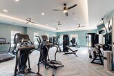Located at 100 Inspire Way in St. Augustine, Florida, this fitness center offers a thoughtfully designed workout environment spread across two exercise rooms on a single floor. The main exercise room features soft teal-painted walls, herringbone-patterned carpet flooring in neutral and aqua tones, and tray ceilings accented by recessed lighting and multiple ceiling fans. Large windows allow natural light to fill the space, creating a bright and welcoming atmosphere. This room is outfitted with a comprehensive selection of strength and cardio equipment, including multi-station cable machines, a full dumbbell rack, weight benches, treadmills, ellipticals, and upright bikes. Wall-mounted mirrors expand the sense of space and allow for form monitoring during workouts. The second exercise room serves as a dedicated studio space, finished with light hardwood-style flooring and crisp white walls accented by tray ceilings with indirect lighting. A large floor-to-ceiling mirror spans one wall, and built-in shelving provides organized storage for accessories. This room accommodates group fitness activities with a row of spin bikes, medicine balls, resistance bands, and stability equipment. A wall-mounted television supports guided workout sessions. Together, the two rooms provide a versatile and well-appointed fitness facility suited for a wide range of exercise routines.