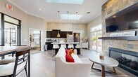 This single-floor community clubhouse is designed to foster a warm, welcoming atmosphere through an open-concept layout that seamlessly connects its key functional zones. The space is anchored by a striking floor-to-ceiling stacked-stone fireplace, topped with a wall-mounted television, which serves as the focal point of the lounge area. Comfortable seating, including a neutral sofa and leather accent chair, creates an inviting gathering space, while large sliding glass doors offer direct access to an outdoor patio furnished with seating and umbrellas.

The kitchen area is outfitted with rich dark cabinetry, light countertops, stainless steel appliances, and a central island with bar-height seating. Pendant lighting and dual skylights flood the space with abundant natural light. A dedicated coffee station adds convenience for daily use.

Adjacent to the main lounge, a casual dining nook features a rustic wood-topped table with mixed metal and upholstered seating, accented by framed artwork and a branded accent wall. A separate office or business center area provides workstations for residents.

The flooring throughout consists of large-format tile with distinctive slate inlay accents, tying the design together cohesively. Crown molding and high ceilings contribute to the polished, community-focused ambiance of the space.