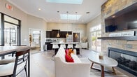 This single-floor community clubhouse is designed to foster a warm, welcoming atmosphere through an open-concept layout that seamlessly connects its key functional zones. The space is anchored by a striking floor-to-ceiling stacked-stone fireplace, topped with a wall-mounted television, which serves as the focal point of the lounge area. Comfortable seating, including a neutral sofa and leather accent chair, creates an inviting gathering space, while large sliding glass doors offer direct access to an outdoor patio furnished with seating and umbrellas.

The kitchen area is outfitted with rich dark cabinetry, light countertops, stainless steel appliances, and a central island with bar-height seating. Pendant lighting and dual skylights flood the space with abundant natural light. A dedicated coffee station adds convenience for daily use.

Adjacent to the main lounge, a casual dining nook features a rustic wood-topped table with mixed metal and upholstered seating, accented by framed artwork and a branded accent wall. A separate office or business center area provides workstations for residents.

The flooring throughout consists of large-format tile with distinctive slate inlay accents, tying the design together cohesively. Crown molding and high ceilings contribute to the polished, community-focused ambiance of the space.
