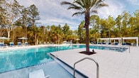 This expansive outdoor amenity space centers around a generously sized swimming pool with crystal-clear blue water, framed by smooth, light-toned tile decking that extends across a broad sundeck. A mature palm tree rises dramatically from a dedicated planter island within the pool area, serving as a natural focal point and adding a distinctly tropical character to the environment.

The surrounding deck is well-appointed with navy blue and white lounge chairs arranged in organized rows, complemented by market umbrellas in teal and white tones that provide shaded relaxation areas. White pergola structures are positioned along the perimeter, offering additional sheltered seating and contributing to the resort-style aesthetic. In-pool sun shelf loungers allow for a more immersive relaxation experience.

A covered outdoor lounge area connects the pool deck directly to the adjacent multi-story building, featuring ceiling fans and comfortable seating beneath an open-air colonnade. Upper balconies overlook the courtyard below. The entire space is bordered by a lush backdrop of tall pines and palms, providing a natural, serene buffer that enhances privacy and reinforces the outdoor ambiance throughout.