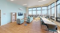 Located at 4051 S 144th St in Tukwila, Washington, this single-floor space is designed to serve as a functional and welcoming community environment. The layout is thoughtfully organized, combining a spacious common lounge area with a fully equipped kitchen and multiple distinct seating zones.

The centerpiece of the space is the expansive common lounge, defined by large, floor-to-ceiling windows along one wall that flood the interior with natural light and provide sweeping outdoor views of the surrounding Tukwila landscape. Light wood flooring runs throughout, complementing the soft aqua and cream wall tones that contribute to a calm, approachable atmosphere. The lounge is furnished with comfortable teal upholstered sofas and armchairs arranged around low pedestal tables, alongside a long communal table with task chairs—supporting both casual gatherings and focused activities. A wall-mounted television and open bookshelves add functional amenities to the space.

The kitchen area features ample cabinetry in a light neutral finish, black appliances including a range, microwave, and full-size refrigerator, and generous counter space. A wall-mounted mini-split system ensures climate control throughout. The overall design prioritizes openness, natural light, and multi-purpose usability.