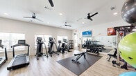 Located in Denham Springs, Louisiana, this fitness center offers a comprehensive workout environment within a single, thoughtfully designed space. The room is finished with light wood-look flooring and crisp white walls, creating a clean and energizing atmosphere throughout. Multiple ceiling fans are mounted overhead, ensuring proper air circulation during workouts, while recessed lighting provides bright, even illumination across the entire floor.

The facility is well-equipped with a diverse range of cardio machines, including treadmills, ellipticals, a recumbent bike, and stationary bikes with integrated digital displays. A tiered dumbbell rack stocked with a full range of free weights anchors the strength training area, accompanied by a multi-function cable machine and an adjustable workout bench. Exercise mats on the floor provide a dedicated space for stretching and floor exercises, while a collection of stability balls in various sizes offers additional functional training options.

Two wall-mounted flat-screen televisions keep members entertained during workouts, and a large mirror along one wall enhances the sense of openness while allowing users to monitor their form. Natural light filters in through multiple windows along the perimeter, connecting the interior to the outdoors. A glass-paneled door provides a secondary access point, contributing to the space's airy, open feel. The overall design is modern, functional, and inviting.