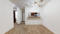 This single-floor residence at 5700 E Mainsgate Rd in Wichita, Kansas offers a straightforward and functional layout throughout. The living area is carpeted and spacious, featuring a ceiling fan with integrated lighting and a textured ceiling. A pass-through counter connects the living space to the kitchen, which is equipped with wood-toned cabinetry, a white electric range, a dishwasher, a refrigerator, and an over-the-range microwave. The kitchen is finished with tile flooring, providing a practical contrast to the carpet found throughout the rest of the home. Multiple bedrooms are present, each carpeted and finished with neutral walls and textured ceilings. One bedroom features a large window fitted with horizontal blinds, allowing natural light to fill the room, while another includes a dark wood-toned door and closet access. A separate living or bonus room includes vertical blinds along a wide window, further enhancing natural light. The bathroom is well-appointed with a single-sink vanity featuring wood cabinetry, a large mirror, tile flooring, and a full tub-shower combination. The overall layout connects seamlessly from room to room, offering a clean and utilitarian living environment suitable for everyday residential use.