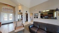 Located at 9257 Somerset Rd in San Antonio, TX, this single-story space exudes a distinct Western character throughout its open, airy layout. The main common area immediately commands attention with dramatically soaring ceilings that amplify the sense of openness, while warm neutral-toned walls provide a welcoming backdrop. Decorative lone star accents are displayed prominently throughout, reinforcing the Texas-inspired aesthetic alongside Western-themed artwork, including a framed horse print and a hat rack near the entrance.

The space features light tile flooring accented by area rugs, and multiple elegant arched doorways connect the central gathering area to adjoining rooms, including what appears to be a dedicated office or study area with built-in shelving. A pass-through kitchen area with dark wood cabinetry is accessible from the main room, offering convenient functionality. An abundance of natural light pours in through a large bank of windows along one wall, offering views of the surrounding outdoor landscape.

The main seating area is furnished with a dark leather loveseat and a patterned accent chair, creating a comfortable and inviting lounge environment. French doors provide an additional point of entry and further enhance the flow of natural light. The overall ambiance is warm, spacious, and distinctly Texan, making this a welcoming and functional single-floor facility.