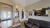 Located at 9257 Somerset Rd in San Antonio, TX, this single-story space exudes a distinct Western character throughout its open, airy layout. The main common area immediately commands attention with dramatically soaring ceilings that amplify the sense of openness, while warm neutral-toned walls provide a welcoming backdrop. Decorative lone star accents are displayed prominently throughout, reinforcing the Texas-inspired aesthetic alongside Western-themed artwork, including a framed horse print and a hat rack near the entrance.

The space features light tile flooring accented by area rugs, and multiple elegant arched doorways connect the central gathering area to adjoining rooms, including what appears to be a dedicated office or study area with built-in shelving. A pass-through kitchen area with dark wood cabinetry is accessible from the main room, offering convenient functionality. An abundance of natural light pours in through a large bank of windows along one wall, offering views of the surrounding outdoor landscape.

The main seating area is furnished with a dark leather loveseat and a patterned accent chair, creating a comfortable and inviting lounge environment. French doors provide an additional point of entry and further enhance the flow of natural light. The overall ambiance is warm, spacious, and distinctly Texan, making this a welcoming and functional single-floor facility.