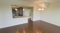 This single-floor residence offers a thoughtfully designed open-concept layout that seamlessly connects the kitchen and living areas, creating a spacious and inviting atmosphere. Rich, dark hardwood flooring runs throughout the main living space, lending warmth and a polished aesthetic to the interior.

The kitchen is a standout feature, equipped with dark espresso-toned cabinetry, stainless steel appliances including a French door refrigerator, and a granite-topped peninsula with an integrated sink. The peninsula provides both additional prep space and a casual seating area, making it ideal for entertaining. Pendant lighting above the peninsula complements the kitchen's modern design.

The adjacent living and dining area benefits from an elegant chandelier with multiple lights, adding a refined touch to the open space. Crown molding along the ceiling adds a classic architectural detail that elevates the overall finish of the home. A tray ceiling detail in the dining area further enhances the sense of dimension and design intention.

Neutral, soft wall tones throughout create a clean and versatile backdrop, allowing the natural character of the flooring and fixtures to take center stage. A hallway visible from the main living area connects to additional rooms, indicating a practical and well-organized floor plan. Overall, the space balances modern finishes with timeless design elements.