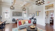 This single-floor community clubhouse is thoughtfully designed to blend comfort and functionality in a bright, inviting atmosphere. The main lounge area serves as the heart of the space, showcasing soaring coffered ceilings, rich hardwood floors, and elegant wainscoting along the walls. Statement pendant lanterns in a matte black finish hang overhead, casting warm light across the room. Comfortable seating, including a plush gray sofa adorned with colorful accent pillows and an orange accent couch, is arranged around a dark round coffee table atop a vibrant multi-toned area rug. A decorative wall installation and framed artwork add personality throughout.

A beverage station tucked along one wall provides a convenient coffee and refreshment setup for occupants. Adjacent to the lounge, a dedicated workspace features a long wooden desk with computer workstations positioned beside tall windows that flood the area with natural light.

A compact kitchenette stands out with bold navy-blue shaker-style cabinetry, white subway tile backsplash, stainless steel appliances, and light countertops—offering a functional and stylish utility area.

A separate meeting or dining room provides a more intimate setting, featuring a round wooden table with upholstered gray chairs, a wall-mounted flat-screen television, wainscoted walls in a soft sage tone, and generous windows overlooking the outdoors.