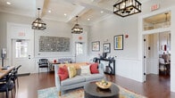 This single-floor community clubhouse is thoughtfully designed to blend comfort and functionality in a bright, inviting atmosphere. The main lounge area serves as the heart of the space, showcasing soaring coffered ceilings, rich hardwood floors, and elegant wainscoting along the walls. Statement pendant lanterns in a matte black finish hang overhead, casting warm light across the room. Comfortable seating, including a plush gray sofa adorned with colorful accent pillows and an orange accent couch, is arranged around a dark round coffee table atop a vibrant multi-toned area rug. A decorative wall installation and framed artwork add personality throughout.

A beverage station tucked along one wall provides a convenient coffee and refreshment setup for occupants. Adjacent to the lounge, a dedicated workspace features a long wooden desk with computer workstations positioned beside tall windows that flood the area with natural light.

A compact kitchenette stands out with bold navy-blue shaker-style cabinetry, white subway tile backsplash, stainless steel appliances, and light countertops—offering a functional and stylish utility area.

A separate meeting or dining room provides a more intimate setting, featuring a round wooden table with upholstered gray chairs, a wall-mounted flat-screen television, wainscoted walls in a soft sage tone, and generous windows overlooking the outdoors.