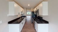 This single-floor space offers a thoughtfully designed layout with a variety of functional rooms finished in a clean, neutral palette. The galley-style kitchen is a standout feature, boasting white shaker cabinetry with matte black hardware, dark granite countertops, a stainless steel sink, and stainless steel appliances including a refrigerator, microwave, and electric range. Wood-look flooring runs through the kitchen and extends into the open living areas, lending warmth and continuity throughout.

The spacious living area benefits from large windows that invite natural light and provide outdoor views, while a built-in closet with bifold doors adds practical storage. Additional rooms feature soft, neutral-toned carpeting and windows with blinds, creating comfortable, versatile spaces suitable for bedrooms or flexible use.

Two full bathrooms serve the space. One includes a tub-shower combination with white tile surround, a dark-stained vanity with a white countertop, and a large mirror. The second bathroom mirrors this style and features a glass-door shower enclosure.

The dedicated laundry room is equipped with a full-size washer and dryer, a utility sink, and wood-look flooring, offering both convenience and practicality in a well-lit, functional setting.