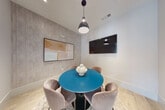 This compact, single-floor meeting room is thoughtfully designed to balance functionality with contemporary style. At its center sits a striking round teal table accompanied by four plush, taupe velvet chairs, creating an inviting and collaborative atmosphere. The flooring is light-toned hardwood, which lends warmth and brightness to the space, complemented by a soft area rug beneath the seating arrangement.

One wall features a textured, stone-look panel finish that adds depth and visual interest, serving as the backdrop for a framed abstract artwork in muted earth tones. A wall-mounted flat-screen display on an adjacent wall ensures the room is equipped for presentations and media viewing. Overhead, a single pendant light with a matte dark fixture provides focused illumination over the table, while recessed lighting supplements the overall brightness.

A sleek, black-framed glass door connects the room to an adjacent, more expansive space, which appears bright and open with large windows overlooking greenery — allowing natural light to filter through and providing a sense of visual connection to the broader environment. The overall ambiance is clean, modern, and professional, making this room well-suited for intimate meetings, focused discussions, or small group work sessions.