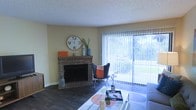 This single-floor living space centers around a charming brick fireplace, which serves as a focal point along one wall and adds a warm, traditional character to the room. The fireplace is framed by a brick hearth and mantel, where decorative accents are displayed. Adjacent to the fireplace, a large sliding glass door with vertical blinds spans nearly the full height of the wall, allowing abundant natural light to flood the interior and offering a view of lush greenery outside. The hard-surface flooring in a rich, dark tone runs throughout the room, providing a grounding contrast to the neutral, light-toned walls. A textured popcorn ceiling adds a classic finish overhead. The living area is furnished with a glass-top coffee table and a butterfly chair, creating a casual, functional seating arrangement. The overall ambiance is bright and comfortable, blending traditional elements like the brick fireplace with lighter, airy qualities provided by the oversized glass door and natural light.