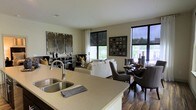 This single-floor space showcases an open-concept layout that seamlessly connects the kitchen, dining, and living areas into one cohesive environment. The kitchen features a generous island with a light stone countertop and a double undermount sink, providing both practical workspace and a natural visual anchor for the open floor plan. Dark hardwood flooring runs throughout, adding warmth and contrast against the neutral, light-toned walls.

The dining area accommodates a dark wood table surrounded by upholstered charcoal chairs, positioned adjacent to large black-framed windows that invite natural light and offer views of the outdoors. Decorative wall art with a grid motif adds a contemporary touch to this zone.

The living area is furnished with a white sectional sofa accented by gold and patterned throw pillows, alongside additional gray accent chairs. A media console and curated wall art panels anchor the space visually. Multiple windows fitted with roller shades and sheer curtains allow for versatile light control while maintaining a bright, airy atmosphere. The overall ambiance is modern and sophisticated, balancing comfort with contemporary design sensibilities.