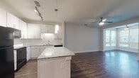 This single-story, one-bedroom, one-bathroom unit offers a well-designed open-concept layout enhanced by dark hardwood-style flooring throughout the main living areas. The kitchen is a standout feature, equipped with white shaker cabinetry, a white subway tile backsplash, granite countertops, black appliances including a refrigerator, range, and built-in microwave, and a central island with additional granite surface space. Pendant lighting and track lighting illuminate the kitchen, while a ceiling fan serves the adjacent living area, which flows seamlessly from the kitchen. A solarium adds a bright, light-filled bonus space with windows that invite natural light. The bedroom is carpeted for comfort, includes a ceiling fan, and receives natural light through double windows with blinds. The bathroom features large-format tile flooring, a granite-topped vanity with white cabinetry, a framed mirror, and a soaking tub with tiled surround. A dedicated laundry area houses a full-sized side-by-side washer and dryer with overhead wire shelving for additional storage. The unit's neutral color palette and modern finishes create a clean, cohesive aesthetic throughout.