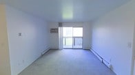 This single-floor space offers a clean and functional layout designed for comfortable everyday living. The main living area is finished with wall-to-wall carpeting that adds warmth underfoot, while bright white walls create a neutral, open atmosphere throughout. A large sliding glass door serves as the room's focal point, flooding the interior with abundant natural light and providing direct access to a private outdoor balcony with a classic railed deck — ideal for enjoying fresh air and outdoor views. Sheer curtains frame the doorway, softening the light that pours in. Climate control is well-considered, with baseboard heating units running along the perimeter walls to ensure even warmth during cooler months, complemented by a wall-mounted air conditioning unit for year-round comfort. The overall ambiance is bright, airy, and move-in ready, with a straightforward design that allows for flexible furnishing and personalization. The space presents a practical and inviting environment, with its connection to the outdoor balcony being a notable highlight that enhances both the sense of space and the quality of natural light within.