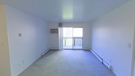 This single-floor space offers a clean and functional layout designed for comfortable everyday living. The main living area is finished with wall-to-wall carpeting that adds warmth underfoot, while bright white walls create a neutral, open atmosphere throughout. A large sliding glass door serves as the room's focal point, flooding the interior with abundant natural light and providing direct access to a private outdoor balcony with a classic railed deck — ideal for enjoying fresh air and outdoor views. Sheer curtains frame the doorway, softening the light that pours in. Climate control is well-considered, with baseboard heating units running along the perimeter walls to ensure even warmth during cooler months, complemented by a wall-mounted air conditioning unit for year-round comfort. The overall ambiance is bright, airy, and move-in ready, with a straightforward design that allows for flexible furnishing and personalization. The space presents a practical and inviting environment, with its connection to the outdoor balcony being a notable highlight that enhances both the sense of space and the quality of natural light within.