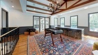 This single-floor space blends classic architectural character with eclectic, modern design sensibilities. Exposed wooden ceiling beams and tray ceilings add warmth and visual depth throughout, while rich hardwood flooring and large, multi-pane windows flood the interior with natural light and offer views of surrounding greenery.

A standout lounge area anchors one section of the space, centered on a full-height brick fireplace with a stone mantel and wall-mounted television, flanked by a pair of plush olive-green velvet armchairs and a marble-topped coffee table. Dark wainscoting lines the lower walls, lending a refined, sophisticated contrast to the bright upper walls.

An adjoining entertainment area serves as a dynamic gathering hub, featuring a foosball table set atop a vibrant, ornately patterned Persian-style area rug. A built-in wet bar with dark cabinetry and gold bar stools lines one wall, while French doors open to additional natural light and outdoor access.

A dramatic bold floral mural in dark tones commands attention near the staircase landing, serving as a striking artistic focal point. Nearby, an arcade gaming station with leather pouf seating adds a playful element. The overall ambiance is sophisticated yet inviting, seamlessly merging traditional architectural details with bold, contemporary décor choices.