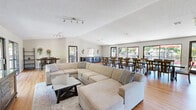 Located at 5016 Paseo Padre Parkway in Fremont, California, this single-floor community space offers a well-designed, open-plan layout that seamlessly connects a spacious living room and a generously sized dining area. Warm hardwood flooring runs throughout, lending a cohesive and inviting feel to the entire space.

The living room is bright and airy, anchored by a large sectional sofa and a media console with a wall-mounted flat-screen television. Track lighting along the textured ceiling provides ample illumination, while large sliding glass doors frame views of a surrounding outdoor deck and lush greenery, allowing natural light to pour in throughout the day.

The dining area is equally impressive, accommodating a long glass-top dining table with seating for a large group — ideal for gatherings and events. Expansive sliding windows and a glass door open directly to the outdoor deck, where views of trees and outdoor seating areas with umbrellas create a pleasant, resort-like atmosphere. A sideboard along one wall offers additional storage and serving space.

The overall ambiance is clean, modern, and functional, with a neutral color palette, textured ceilings, and an open layout that promotes a sense of spaciousness. This community space is well-suited for social events, meetings, or casual gatherings in the heart of Fremont.