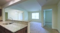 This single-floor space showcases an open-concept design that connects the kitchen and living areas into one cohesive layout. The kitchen features striking granite countertops with warm brown cabinetry, offering generous prep and serving space. A half-wall peninsula separates the kitchen from the adjoining dining area, which is illuminated by a chandelier, creating a defined yet open dining nook.

The living area is finished with plush carpeting and painted in soft, neutral tones that contribute to a bright and welcoming ambiance. A centered window allows natural light to fill the space, enhancing the airy feel of the room. The smooth ceilings feature subtle tray-style detailing near the kitchen transition, adding a touch of architectural interest.

A hallway visible from the living area leads to additional rooms, suggesting a thoughtful floor plan with clear separation between living and private spaces. Overall, the space presents a clean, move-in-ready aesthetic with a harmonious blend of functional design and comfortable finishes throughout.