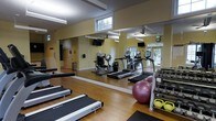 This fitness center is designed to support a comprehensive workout experience within a single-floor layout. The space features warm yellow walls, wood-style flooring, and large floor-to-ceiling mirrors along one wall, which enhance the sense of openness and allow users to monitor their form during exercise. Natural light filters in through multiple windows positioned along the walls, contributing to a bright and energizing atmosphere, while ceiling fans and overhead fluorescent lighting provide additional comfort and visibility.

The cardio section is outfitted with multiple TRUE-brand treadmills, elliptical trainers, and a stationary bike, each equipped with individual digital display consoles. A wall-mounted television is positioned for viewing during workouts. The strength training area includes a multi-functional cable machine with an adjustable bench, catering to a variety of resistance exercises. A dedicated free weights section features a dumbbell rack stocked with kettlebells and hand weights, along with a stability ball for core training. Rubber flooring mats are placed beneath the strength equipment for safety. The overall layout is organized and functional, offering a well-rounded fitness environment.