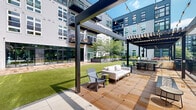 Located at 101 Washington St in Conshohocken, Pennsylvania, this thoughtfully designed outdoor amenity space offers a compelling blend of modern architecture and natural landscaping. The centerpiece of the space is a generous, well-manicured lawn area framed by raised stone planters filled with ornamental grasses and shrubs, creating a lush, park-like atmosphere within the courtyard setting. Seating areas with wooden chairs and tables are arranged along the lawn's perimeter, providing comfortable spaces for gathering and relaxation. A striking pergola structure anchors one end of the outdoor space, outfitted with bold red hanging swing seats and wood-tiled flooring, offering a shaded retreat with a distinctly social ambiance. Adjacent to the pergola, an elevated terrace with a metal railing system provides panoramic views of the surrounding tree canopy and neighboring office buildings, reinforcing the property's connection to its green surroundings. The multi-story building facades feature a mix of brick, fiber cement siding, and large windows, while private balconies overlook the courtyard below. Interior access points with glass-panel doors connect the outdoor areas to the building's interior. The overall design prioritizes open-air living, community interaction, and seamless integration between the built environment and its natural surroundings.
