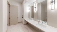 Located at 558 E Stonewall St in uptown Charlotte, NC, this single-floor commercial facility is thoughtfully designed to provide a clean, functional, and well-appointed experience. The space is finished throughout with large-format light tile flooring that lends a bright, cohesive aesthetic.

The vanity area features a continuous countertop with multiple undermount sinks, wall-mounted sconce lighting, and large mirrors that maximize both visibility and the sense of openness. Clean white walls and crisp fixtures maintain a polished, professional appearance throughout.

A dedicated shower suite offers a spacious, accessible walk-in shower with textured mosaic stone tile flooring, a handheld showerhead with an adjustable slide bar, grab bars for safety, and white subway-style tile walls. A bamboo bath mat adds a warm, natural touch to the otherwise clean, minimal design.

A separate lounge or changing area provides a comfortable retreat, featuring a wall-mounted flat-screen television, a bold geometric-patterned accent wall in deep tones, built-in cabinetry for storage, a cushioned bench, and a decorative area rug — together creating a distinctly refined atmosphere within a functional setting.

The men's restroom area includes private urinal stalls separated by clean white partition walls, consistent with the facility's overall neutral and utilitarian design language. This facility is well-suited for a gym, athletic club, or upscale commercial venue.
