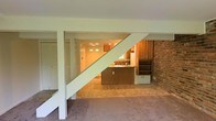 This two-floor space offers a functional and open layout that connects its main living areas in a straightforward, practical design. The main level features a carpeted living room that draws immediate attention with its large sliding glass doors, which flood the room with natural light and provide a direct view of a lush green outdoor area. The bright, airy feel of this space is enhanced by clean white walls and generous ceiling height.

Adjacent to the living room, the kitchen is defined by warm wood cabinetry and a central island that serves as a natural dividing point between the cooking and living areas. A notable brick accent wall adds a touch of character and texture to the space, complementing the earthy tones of the cabinetry and tiled flooring. A staircase with white painted railings leads to the second floor, positioned to maintain an open sightline between the main living area and the kitchen. Small transom-style windows near the kitchen entry allow additional natural light to filter through. Overall, the space presents a classic, no-frills interior with practical features and inviting natural light throughout.