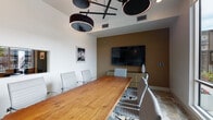 This thoughtfully designed single-floor space blends functionality with contemporary style, offering distinct areas suited for both professional and social use. The conference room serves as a standout feature, centered around a solid wood live-edge table paired with sleek white ergonomic chairs. A large wall-mounted display screen is set against a warm earth-toned accent wall, while a striking multi-drum pendant light fixture overhead adds a bold design element. A geometric-patterned floor tile grounds the room, and large windows invite natural light and views of the surrounding outdoor area. A full-height glass partition wall separates the conference room from the adjacent open lounge and kitchen area, maintaining visual connectivity throughout the space. The open-plan lounge features light hardwood flooring, a round dining table with vibrant orange upholstered chairs, and a curated arrangement of shelving and décor. The kitchen is anchored by a large marble-topped island with green bar seating, complemented by modern pendant lighting and sleek cabinetry. Floor-to-ceiling glass doors open to an outdoor area filled with natural greenery, flooding the interior with abundant daylight and creating a seamless indoor-outdoor connection.