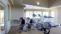 This fitness center offers a practical and well-organized space designed to support a range of cardiovascular workouts. The room is outfitted with multiple cardio machines, including upright stationary bikes, elliptical trainers, and treadmills, providing ample options for users of varying fitness levels and preferences. The equipment is arranged to allow comfortable spacing between machines, promoting an open and accessible workout environment. A wall-mounted water fountain is conveniently positioned near the entrance, ensuring hydration is easily accessible during exercise sessions. Natural light flows in through a row of large windows along one wall, offering views of the surrounding exterior and creating a bright, pleasant atmosphere. The ceiling features recessed lighting supplemented by a wall-mounted fixture, ensuring the space remains well-lit at any time of day. Carpeted flooring covers most of the room, contributing to a quieter, cushioned feel underfoot. A wall-mounted television is positioned for visibility from the cardio equipment, adding an entertainment element to workouts. The overall ambiance is clean, functional, and inviting, making this fitness center a convenient and effective amenity for its users.