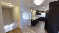 This single-floor residence offers a thoughtfully designed open-concept layout that seamlessly connects the kitchen and living areas, creating a spacious and airy atmosphere. The kitchen is a standout feature, showcasing sleek dark cabinetry that contrasts beautifully with light granite countertops. A central island provides additional workspace and serves as a natural gathering point, complete with pendant lighting that adds a warm, modern ambiance. Additional track lighting overhead ensures the space is well-lit and functional. The kitchen also features a tile backsplash with a decorative accent detail, adding a touch of character to the space. Hardwood-style flooring runs throughout the open living area, providing a warm and cohesive aesthetic. The living area is generously proportioned, with clean white walls and high ceilings that enhance the sense of openness. Natural light filters into the space, contributing to its bright and welcoming feel. The overall design balances contemporary style with practical functionality, making it an inviting and versatile living environment.