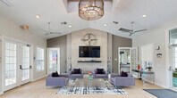 Located at 17800 Murdock Cir in Port Charlotte, Florida, this single-story community clubhouse offers a well-appointed gathering space designed to serve residents across multiple functional areas. The centerpiece of the facility is a dramatic open lounge featuring soaring vaulted ceilings, large-format tile flooring, and an eye-catching floor-to-ceiling tiled accent wall adorned with a decorative metal compass sculpture and a wall-mounted flat-screen TV. Multiple sets of French doors line the lounge, inviting abundant natural light and providing easy access to the outdoors. Comfortable gray upholstered seating is arranged around a central coffee table atop a graphic area rug, creating an inviting social atmosphere. A ceiling skylight and modern drum chandelier add architectural character overhead. The fitness center is well-equipped with cardio machines, free weights, resistance bands, exercise balls, and full-wall mirrors, all set against bold navy blue accent walls. The business center offers wood-look flooring, soft sage-green walls, desktop computers, a printer station, and ample seating for productivity. A fully equipped kitchen with dark cabinetry, white countertops, stainless steel appliances, and dual windows rounds out this versatile community space.