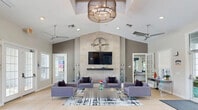 Located at 17800 Murdock Cir in Port Charlotte, Florida, this single-story community clubhouse offers a well-appointed gathering space designed to serve residents across multiple functional areas. The centerpiece of the facility is a dramatic open lounge featuring soaring vaulted ceilings, large-format tile flooring, and an eye-catching floor-to-ceiling tiled accent wall adorned with a decorative metal compass sculpture and a wall-mounted flat-screen TV. Multiple sets of French doors line the lounge, inviting abundant natural light and providing easy access to the outdoors. Comfortable gray upholstered seating is arranged around a central coffee table atop a graphic area rug, creating an inviting social atmosphere. A ceiling skylight and modern drum chandelier add architectural character overhead. The fitness center is well-equipped with cardio machines, free weights, resistance bands, exercise balls, and full-wall mirrors, all set against bold navy blue accent walls. The business center offers wood-look flooring, soft sage-green walls, desktop computers, a printer station, and ample seating for productivity. A fully equipped kitchen with dark cabinetry, white countertops, stainless steel appliances, and dual windows rounds out this versatile community space.