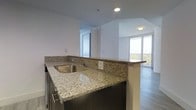This single-floor space showcases a thoughtfully designed open-concept layout that seamlessly connects the kitchen and living areas. The kitchen is appointed with speckled granite countertops that extend into a peninsula-style breakfast bar, complete with a stainless steel undermount sink and dark espresso-toned cabinetry with sleek hardware. Recessed lighting illuminates the kitchen area, highlighting the clean lines of the countertop surfaces.

The open layout transitions naturally toward the living area, where large floor-to-ceiling windows and a sliding glass door invite abundant natural light throughout the space. The light-toned hardwood-style flooring runs consistently across both areas, contributing to a cohesive and contemporary aesthetic. The white walls enhance the brightness of the interior, while the subtle tray ceiling detail near the living area adds a touch of architectural interest. The expansive glazing provides views of the surrounding exterior, making the living space feel open and airy. Overall, the unit presents a clean, modern aesthetic well-suited for comfortable everyday living.
