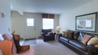 This two-story residential space offers a practical and comfortable layout designed for everyday living. The main level begins with an inviting living room, where a front entry door with decorative glass panels welcomes residents and guests. Natural light filters in through a window dressed with a dark valance, brightening the carpeted space. The living room is well-proportioned, providing ample room for seating arrangements, as seen with the dark leather sofa and matching armchair that anchor the area. Neutral-toned walls and light carpet create a calm, understated ambiance throughout. The overall design is straightforward and functional, with clean lines and a layout that maximizes usable living space. The two-floor configuration suggests additional rooms above, likely accommodating bedrooms and further living areas, offering a clear separation between common living spaces on the main level and private spaces on the upper floor. The home's interior reflects a modest, livable aesthetic with practical finishes that provide a solid foundation for personalization and comfortable day-to-day use.