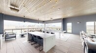 This rooftop amenity space offers a thoughtfully designed open-plan layout that blends modern aesthetics with a relaxed, social atmosphere. The interior is anchored by a whitewashed wood-plank ceiling adorned with warm Edison string lights, creating an inviting ambiance throughout. Light wood-toned tile flooring runs seamlessly across the entire floor, complementing the cool gray wall tones and textured brick accent wall.

A central white marble-topped island with bar-height seating serves as a natural gathering point, while a sleek white pool table positioned on a patterned area rug provides a focal point for recreation. A dedicated lounge area features comfortable upholstered seating arranged around a wall-mounted flat-screen television set against the white brick accent wall, with a large ceiling fan overhead.

The bar area, finished with striking teal subway tile, includes counter seating and shelving, making it an ideal entertainment hub. Expansive floor-to-ceiling windows and open-air balcony access on multiple sides flood the space with natural light and frame sweeping panoramic views of the surrounding cityscape. The outdoor balcony, fitted with glass railings and bistro seating, extends the entertaining experience with unobstructed skyline vistas. Ceiling fans throughout ensure comfort, completing this versatile and well-appointed communal retreat.