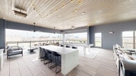 This rooftop amenity space offers a thoughtfully designed open-plan layout that blends modern aesthetics with a relaxed, social atmosphere. The interior is anchored by a whitewashed wood-plank ceiling adorned with warm Edison string lights, creating an inviting ambiance throughout. Light wood-toned tile flooring runs seamlessly across the entire floor, complementing the cool gray wall tones and textured brick accent wall.

A central white marble-topped island with bar-height seating serves as a natural gathering point, while a sleek white pool table positioned on a patterned area rug provides a focal point for recreation. A dedicated lounge area features comfortable upholstered seating arranged around a wall-mounted flat-screen television set against the white brick accent wall, with a large ceiling fan overhead.

The bar area, finished with striking teal subway tile, includes counter seating and shelving, making it an ideal entertainment hub. Expansive floor-to-ceiling windows and open-air balcony access on multiple sides flood the space with natural light and frame sweeping panoramic views of the surrounding cityscape. The outdoor balcony, fitted with glass railings and bistro seating, extends the entertaining experience with unobstructed skyline vistas. Ceiling fans throughout ensure comfort, completing this versatile and well-appointed communal retreat.