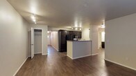 This single-floor space is designed with an open-concept layout that seamlessly connects the main living area to the kitchen. The kitchen is outfitted with dark espresso cabinetry, black appliances including a refrigerator, microwave, and range, and a peninsula-style breakfast bar that doubles as a natural divider between the kitchen and the living space. Track lighting runs along the ceiling, providing adjustable illumination across the main area and complementing the recessed lighting fixtures found throughout.

The flooring is consistent throughout the space, featuring rich, dark hardwood-style planks that add warmth and a cohesive visual flow from room to room. The neutral, light-toned walls contrast well with the darker finishes, keeping the space feeling open and balanced.

A closet with a rod and shelving is visible near the entrance, offering dedicated storage. A hallway extends toward additional rooms, with a glimpse of what appears to be a bathroom visible in the background. The overall layout is practical and well-organized, with clearly defined zones for living, cooking, and storage, making efficient use of the available space.