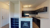 This fully renovated one-bedroom, one-bathroom residence offers a thoughtfully updated interior across a single floor. The open-concept living and kitchen area serves as the centerpiece of the space, anchored by a distinctive tiled fireplace with built-in open shelving that separates the living area from the kitchen. The kitchen has been renovated with dark cabinetry featuring sleek hardware, light granite countertops, wood-look flooring, and black appliances including a refrigerator, range, and dishwasher. A stainless steel double-basin sink completes the functional layout, while a track lighting fixture provides focused illumination over the workspace.

The bedroom is generously proportioned, featuring plush wall-to-wall carpeting and multiple windows fitted with horizontal blinds that allow abundant natural light. The full bathroom has been updated with wood-look vinyl flooring, an extended vanity with a light countertop and integrated sink, a large mirror, and a tub-shower combination. The overall color palette throughout the unit is neutral and cohesive, with light walls and warm-toned finishes creating a clean, move-in-ready atmosphere.