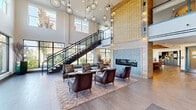 This two-story amenity space makes an immediate impression through its soaring double-height lobby, where floor-to-ceiling windows flood the interior with abundant natural light. A sleek open-riser staircase with black steel framing and glass railings connects both floors, serving as a striking architectural centerpiece. A large-scale textured tile fireplace wall anchors the main lounge area, where leather club chairs and pendant globe lighting create a refined, welcoming atmosphere.

The ground floor extends into a co-working area featuring a substantial solid wood communal desk beneath a reclaimed wood ceiling with exposed beam detailing and an industrial-style chandelier. Nearby, a dedicated conference room offers a metal-topped meeting table, wall-mounted display, and built-in grey cabinetry. A kitchenette with grey cabinetry and stainless steel appliances provides added convenience adjacent to the conference room.

The second floor presents a comfortable lounge with hardwood-style flooring, a large wall-mounted television set within a bold chevron-patterned wood surround, and a bistro-style seating nook with decorative wallcovering. French doors open onto a private balcony, offering outdoor views of the surrounding area.