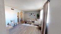 This single-floor residence offers a cohesive, neutral aesthetic throughout, combining light gray walls, wood-look flooring, and textured ceilings to create a warm and inviting atmosphere. The living room serves as the heart of the home, featuring large windows dressed with layered curtains that allow ample natural light to fill the space. Distinctive arched entryways connect the living area to the dining room, adding an architectural charm that sets the home apart. The dining area is intimate and welcoming, with textured walls and a framed art piece that complement the natural wood tones of the dining table. The kitchen is well-appointed with dark espresso cabinetry, light countertops, tile flooring, and black appliances including a range and refrigerator, along with a window that brings in additional natural light. The bedrooms are calm and restful, each featuring wood-look flooring, large windows with soft curtains, and ceiling fans for year-round comfort. The primary bedroom offers generous proportions and a serene ambiance. The bathroom includes a full tub and shower enclosure with clean, simple finishes. Overall, the home presents a cohesive and thoughtfully designed single-level layout ideal for comfortable everyday living.