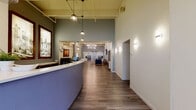 This single-floor lobby and common area presents a thoughtfully designed, contemporary space that balances professional functionality with inviting comfort. The entry immediately draws attention to a sleek, curved reception desk finished in a muted blue-purple tone and topped with a white countertop, accented by pendant lighting that hangs from high, exposed ceilings with industrial undertones.

The open-plan layout flows seamlessly from the reception area into multiple distinct lounge zones, each furnished with mid-century modern seating in neutral and navy tones. A standout lounge features a large-scale photograph of oranges as a bold focal point, complemented by warm wood-toned coffee tables and an eclectic starburst clock. A separate seating alcove centers around a wall-mounted flat-screen TV, with plush sofas and a marble-top round coffee table arranged for a refined, comfortable experience.

Wide-plank wood-look flooring runs throughout, tying the space together with warmth and continuity. The elevator lobby is clean and well-appointed, featuring two stainless steel elevator doors flanked by potted fiddle-leaf fig trees and a cheerful yellow accent door. Curated artwork, vibrant orange typographic signage, and carefully placed greenery contribute to a cohesive, lively aesthetic that feels both modern and welcoming.