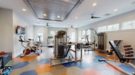 Located at 645 Old Hickory Boulevard in Nashville, TN, this fitness center offers a thoughtfully designed exercise space that balances functionality with visual appeal. The room's most distinctive feature is its eye-catching floor, finished in a bold hexagonal tile pattern of orange, blue, and gray — adding a dynamic, energetic aesthetic to the workout environment.

The space is generously outfitted with a comprehensive selection of Precor strength training machines, including cable and weight systems, as well as a dedicated free weights area with a dumbbell rack and adjustable bench. Cardio equipment includes multiple treadmills, elliptical machines, stationary bikes, and a rowing machine. A wall-mounted television accompanies the cardio section, enhancing the workout experience.

Architectural details elevate the space beyond a typical gym. A tray ceiling with warm wood paneling serves as a striking focal point, complemented by recessed lighting and multiple ceiling fans that promote airflow. Large windows and clerestory windows on multiple walls flood the interior with natural light and offer views of the surrounding greenery. Full-length mirrors along one wall visually expand the room and support proper form during exercise. The overall ambiance is bright, modern, and motivating — a well-appointed fitness facility suited for a wide range of training goals.