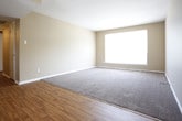 This single-floor space offers a clean, functional layout designed for comfortable everyday living. The main living area is a standout feature, combining warm hardwood flooring at the entryway with plush carpeting throughout the central living zone, creating a welcoming transition between spaces. Neutral beige walls provide a versatile backdrop that suits a wide range of furnishing styles and décor preferences. A large picture window dominates one wall of the living area, flooding the room with abundant natural light and offering views of the surrounding exterior. White baseboards and trim offer a crisp contrast to the warm tones of the flooring and walls, contributing to the space's clean and tidy aesthetic. The open layout allows for flexible furniture arrangement, making the space adaptable to various needs. Overall, the space presents a straightforward, well-maintained interior with a bright and airy ambiance, well-suited for those seeking a simple yet comfortable living environment.