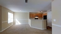 This single-floor residence offers an open-concept design that effortlessly connects the living and kitchen areas into one cohesive, functional space. The living area is finished with plush wall-to-wall carpeting in a warm neutral tone, creating a comfortable and inviting atmosphere. Large windows allow generous natural light to pour in, brightening the space and enhancing its airy feel. Smooth, light-colored walls with classic white baseboards provide a clean and timeless backdrop throughout.

The kitchen is defined by warm honey-toned wooden cabinetry that provides ample storage space. A raised breakfast bar peninsula separates the kitchen from the living area, offering a practical surface for casual dining or meal prep. Black appliances, including a refrigerator and range, add a bold contrast to the lighter surroundings. A track lighting fixture overhead ensures the kitchen is well-lit for everyday use, while a recessed vent hood over the range adds a functional touch. The kitchen also features a built-in sink along the peninsula, contributing to its practical, well-organized layout. Recessed ceiling lighting complements the overall illumination of the space. The combination of neutral tones, natural light, and open flow makes this a welcoming and versatile living environment.