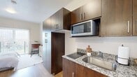 Located at 940 W North Temple St in Salt Lake City, Utah, this efficiently designed one-bedroom, one-bathroom unit offers a thoughtful open-concept layout that maximizes every inch of its compact footprint. Light oak-toned hardwood flooring runs throughout, creating a warm, cohesive feel across all areas of the space.

The kitchen is outfitted with rich dark-wood cabinetry extending to the ceiling for ample storage, granite countertops, a flush cooktop, stainless steel sink, and a built-in microwave — delivering a modern, functional cooking area. The kitchen flows seamlessly into a dining nook and living area, where a small dining table and warm-toned furnishings create an inviting atmosphere.

The bedroom occupies the primary portion of the unit and features a large sliding glass door dressed with vertical blinds, allowing abundant natural light while offering views of the surrounding neighborhood. The room comfortably accommodates a full-sized bed and a workspace desk.

The bathroom is clean and straightforward, featuring a wall-mounted sink, standard toilet, and the same warm hardwood flooring found throughout the unit. A hallway entry connects all areas efficiently, with neutral white walls and recessed lighting maintaining a bright, airy ambiance throughout this well-appointed Salt Lake City residence.