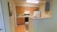 This single-floor space centers around a well-organized kitchen that serves as a focal point of the layout. The kitchen is outfitted with warm-toned wood cabinetry spanning both upper and lower sections, providing generous storage throughout. A white electric range and matching full-size refrigerator supply all essential cooking and food storage needs, while the under-cabinet range hood adds practical ventilation. The countertops are finished in a light, neutral tone that complements the cabinetry and keeps the space feeling bright and cohesive. A peninsula counter extends into the adjoining area, creating a natural breakfast bar that doubles as a casual dining or workspace surface. The flooring throughout the kitchen is light hardwood, contributing warmth to the overall aesthetic. A striped area rug adds a subtle decorative touch at the kitchen entrance. Overhead fluorescent lighting ensures the workspace is well-lit. A hallway visible from the kitchen suggests additional rooms beyond, reinforcing the functional, single-level flow of the space. The overall ambiance is clean, practical, and straightforward.
