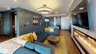 This thoughtfully designed single-floor space blends contemporary aesthetics with warm, inviting details throughout. The main lounge area serves as the centerpiece, anchored by a sleek linear fireplace set against a marble-style surround with a wall-mounted television above. A striking tiered crystal chandelier commands attention overhead, casting a warm glow across plush teal sectional sofas accented with mustard yellow throw pillows. Rich hardwood flooring runs throughout, complemented by a bold patterned area rug. Textured, deep-toned feature walls add depth and sophistication to the lounge, while curated artwork contributes to the space's artistic character.

The kitchen is equally impressive, featuring exposed wood ceiling beams, light wood cabinetry, clean white countertops, and a gas cooktop island. Decorative teal patterned tile walls provide a vibrant focal point, and stylish pendant lighting in copper and matte black tones adds an industrial-modern touch. The kitchen is fully equipped for serious culinary use.

Adjacent to the lounge, the dining area offers seating for a large group around a stunning live-edge wood table, illuminated by sculptural white pendant lights. Floor-to-ceiling sliding glass doors open to an outdoor deck, flooding the space with natural light and offering expansive city skyline views.